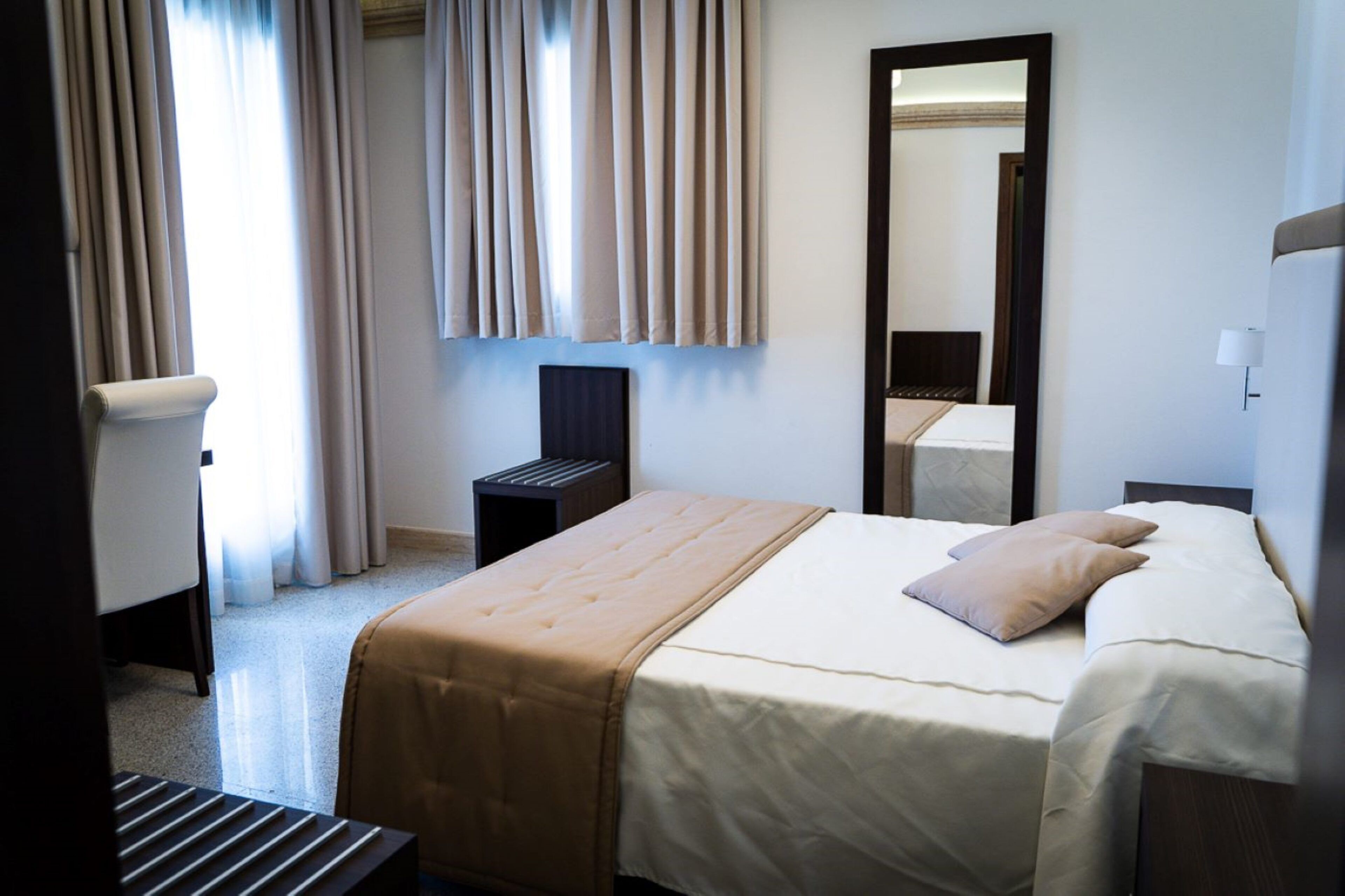 A hotel room with a bed, a chair, a bedside table, a mirror, and curtains.
