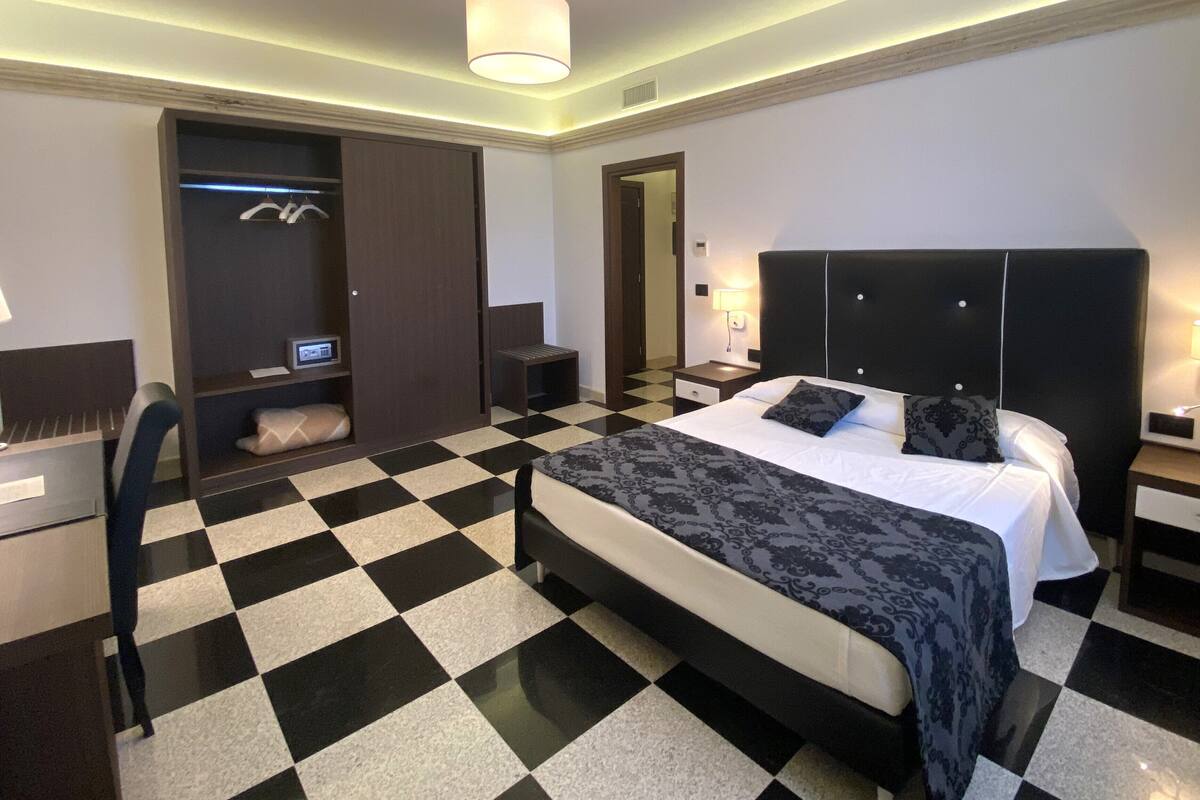 Una habitación de hotel con piso a cuadros, una cama grande con cabecera negra, un armario, un escritorio y una mesita de noche con lámpara.
