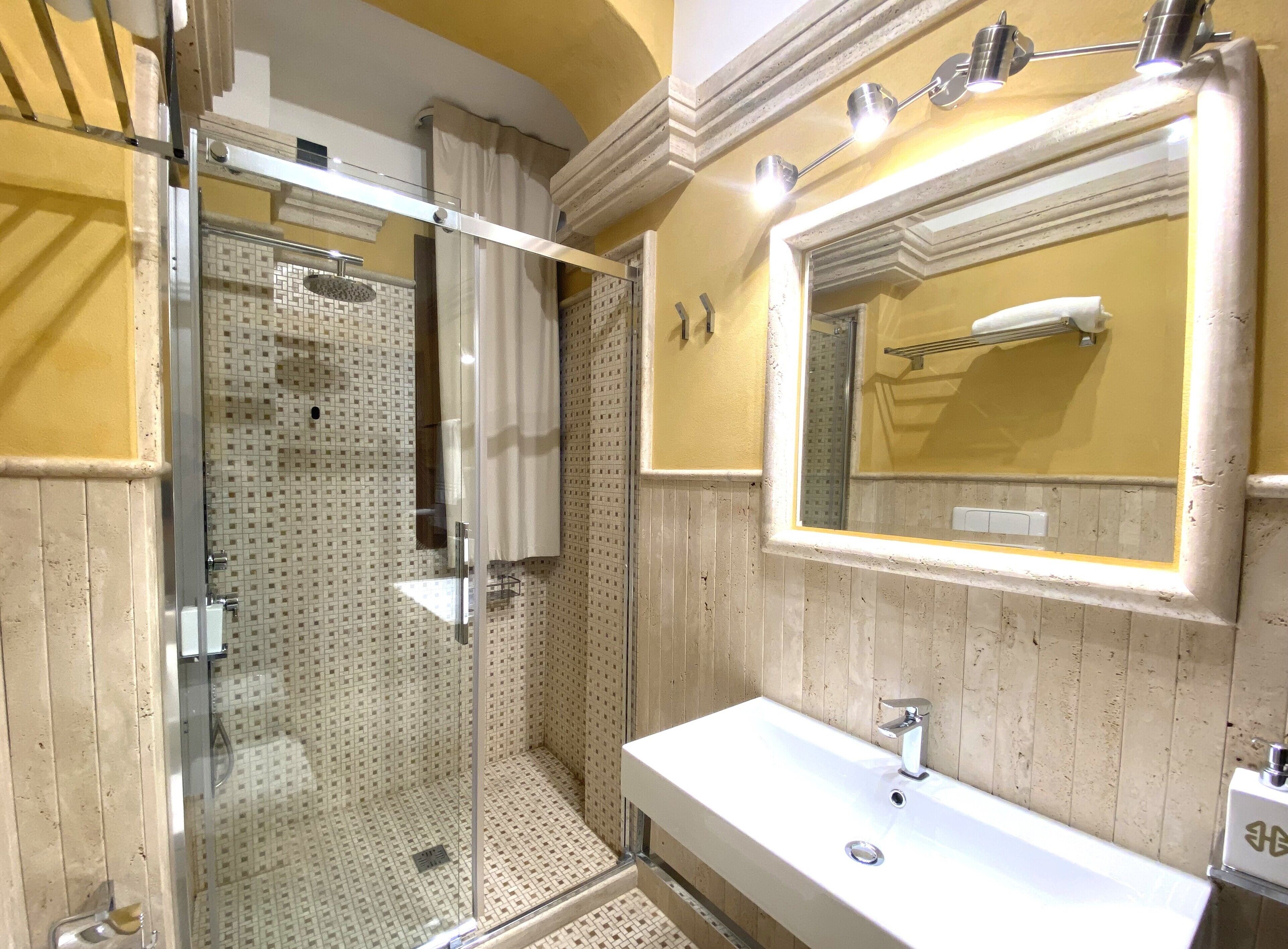 Un baño con ducha de vidrio, un lavamanos blanco y un espejo grande con luces arriba.