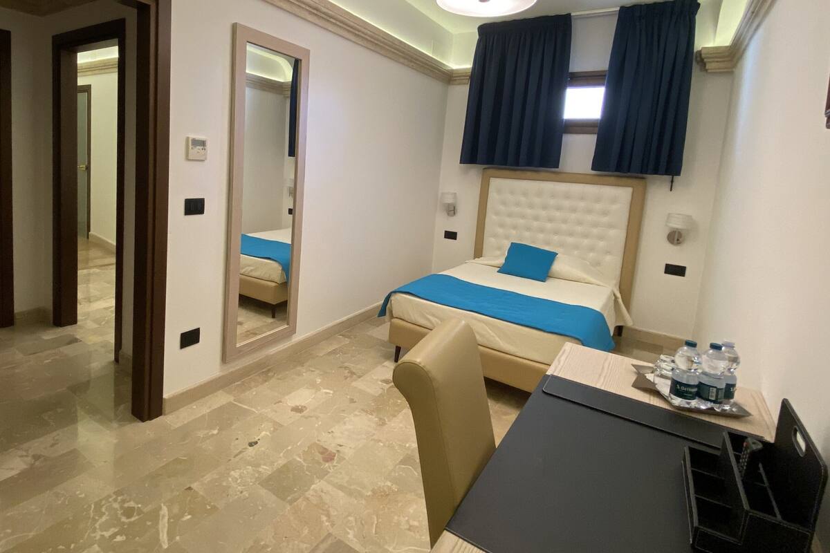 Habitación de hotel con cama, escritorio, silla, espejo, ventana con cortinas y un porta botellas de agua.