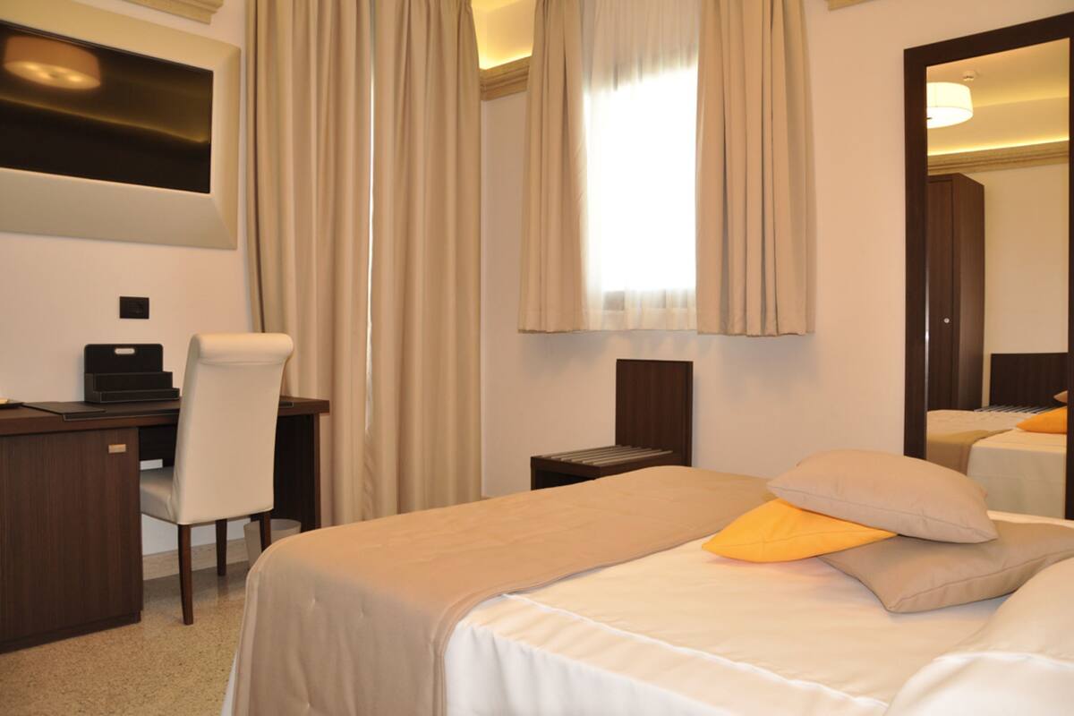 Habitación de hotel con cama, escritorio, silla, espejo y ventana con cortinas.