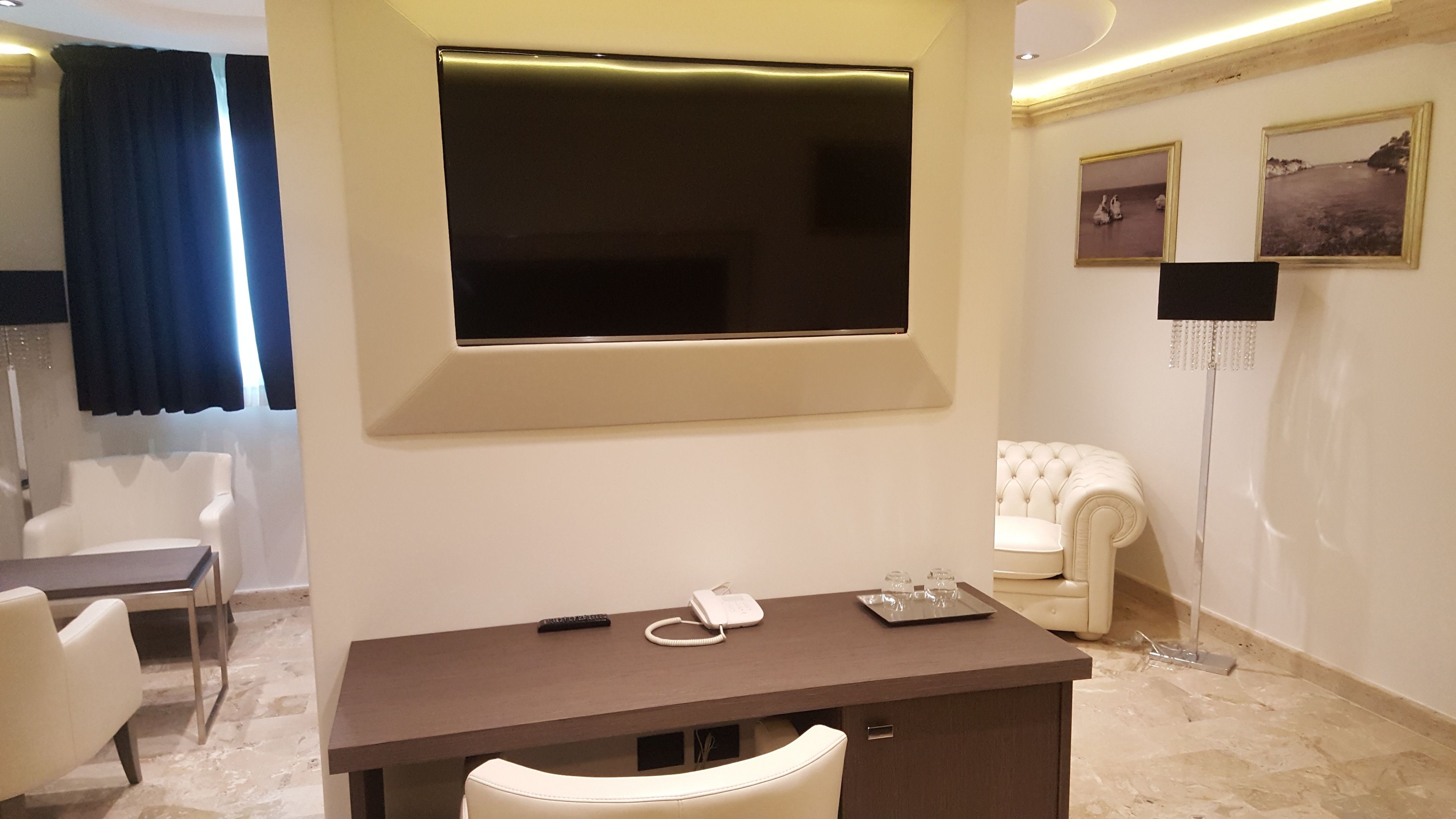 Habitación de hotel con un televisor de pantalla plana grande montado en la pared, un escritorio con teléfono y control remoto, un sillón de cuero blanco y una lámpara de pie.