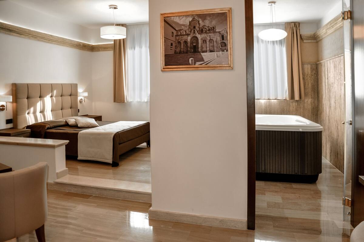 Una habitación de hotel con una cama grande, una zona de estar con una mesa y sillas, una bañera y un cuadro en la pared.