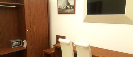 A room with a wooden desk, two chairs, a small table, a wardrobe, and a framed picture on the wall.
