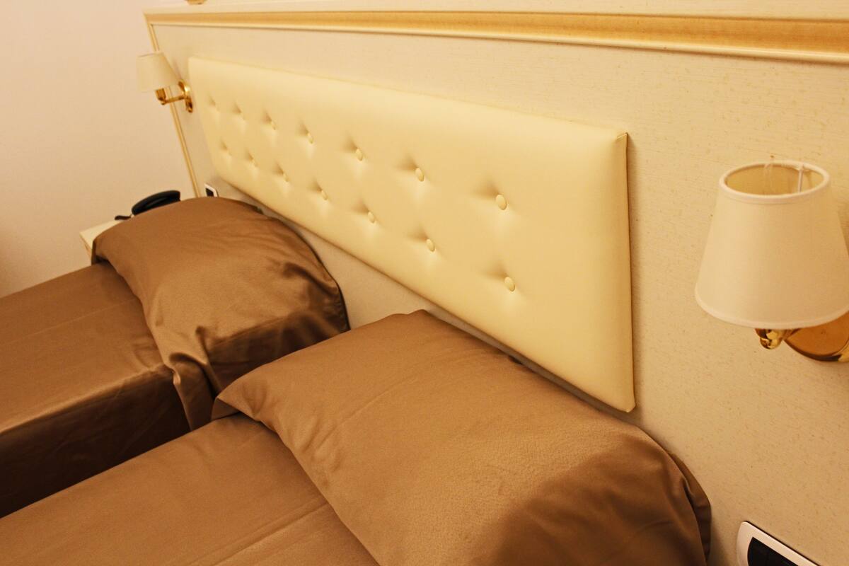 A hotel room with a tufted headboard, two beds with brown bedding, a bedside lamp, and a wall-mounted light fixture.