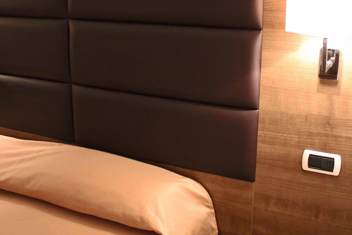 A hotel room with a padded headboard, a bedside table with a lamp, and a small bedside table with a phone.