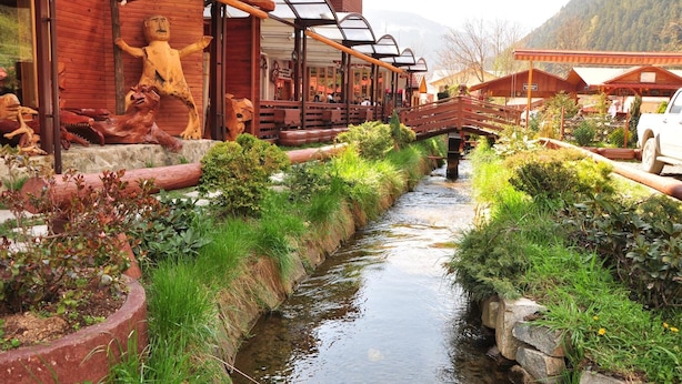 A wooden building with a covered outdoor seating area, a narrow water channel, and a white truck parked nearby. - Inan Kardesler Hotel (Çaykara)