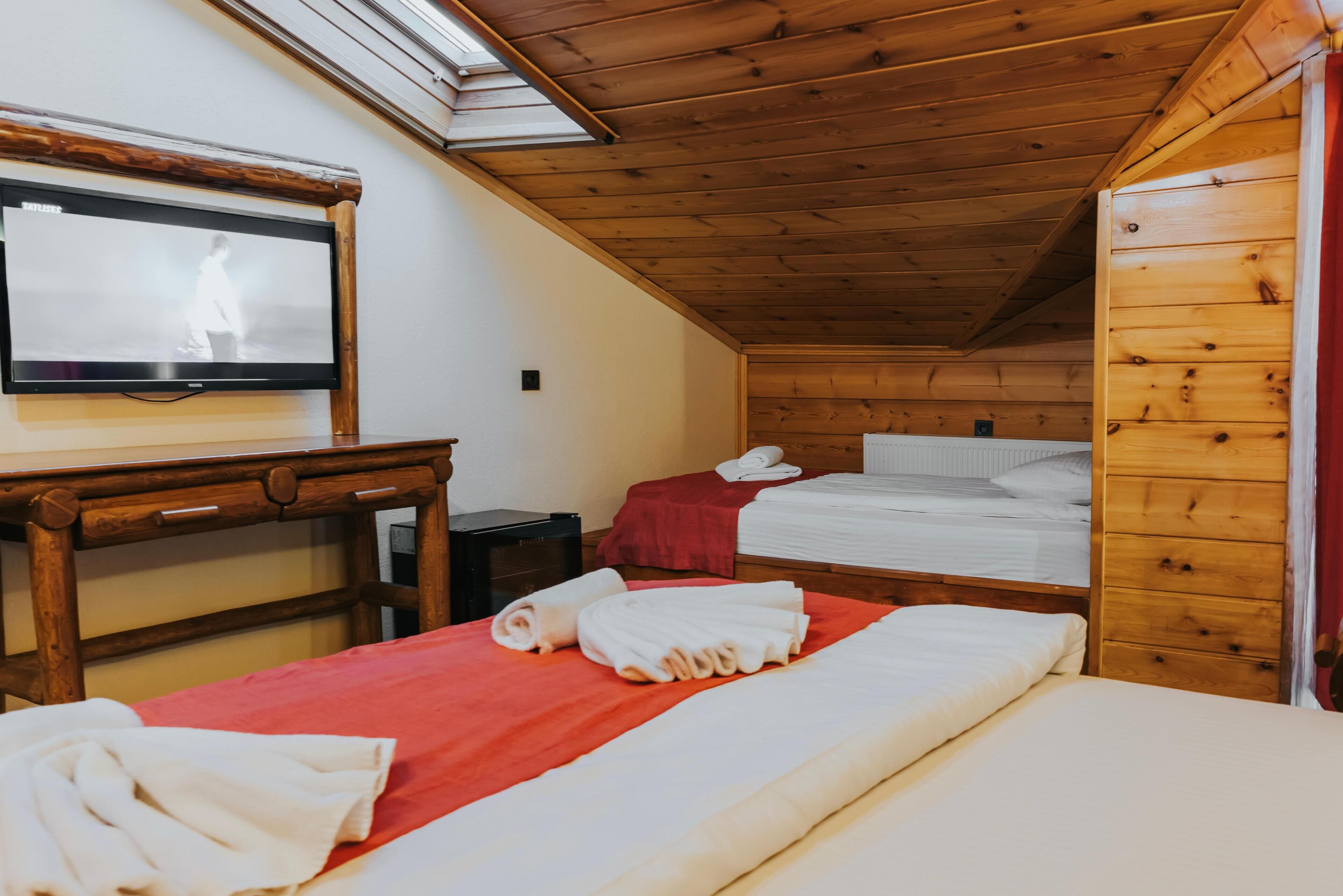 A room with a wooden ceiling, a TV mounted on the wall, two beds with red and white bedding, and a wooden dresser.