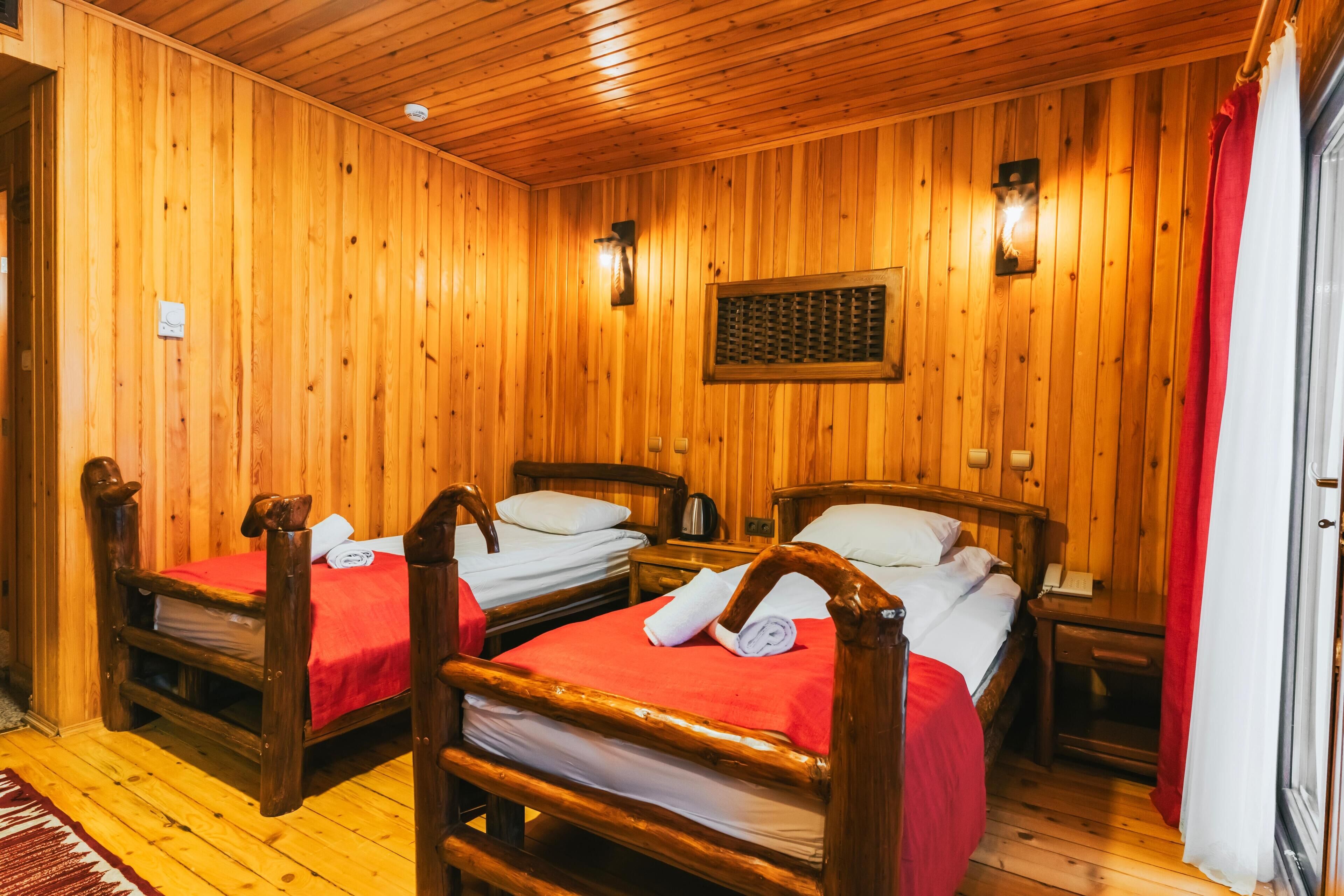 A wooden room with two beds, each with a red bedspread and white pillows, wooden headboards, and a wooden wall panel.