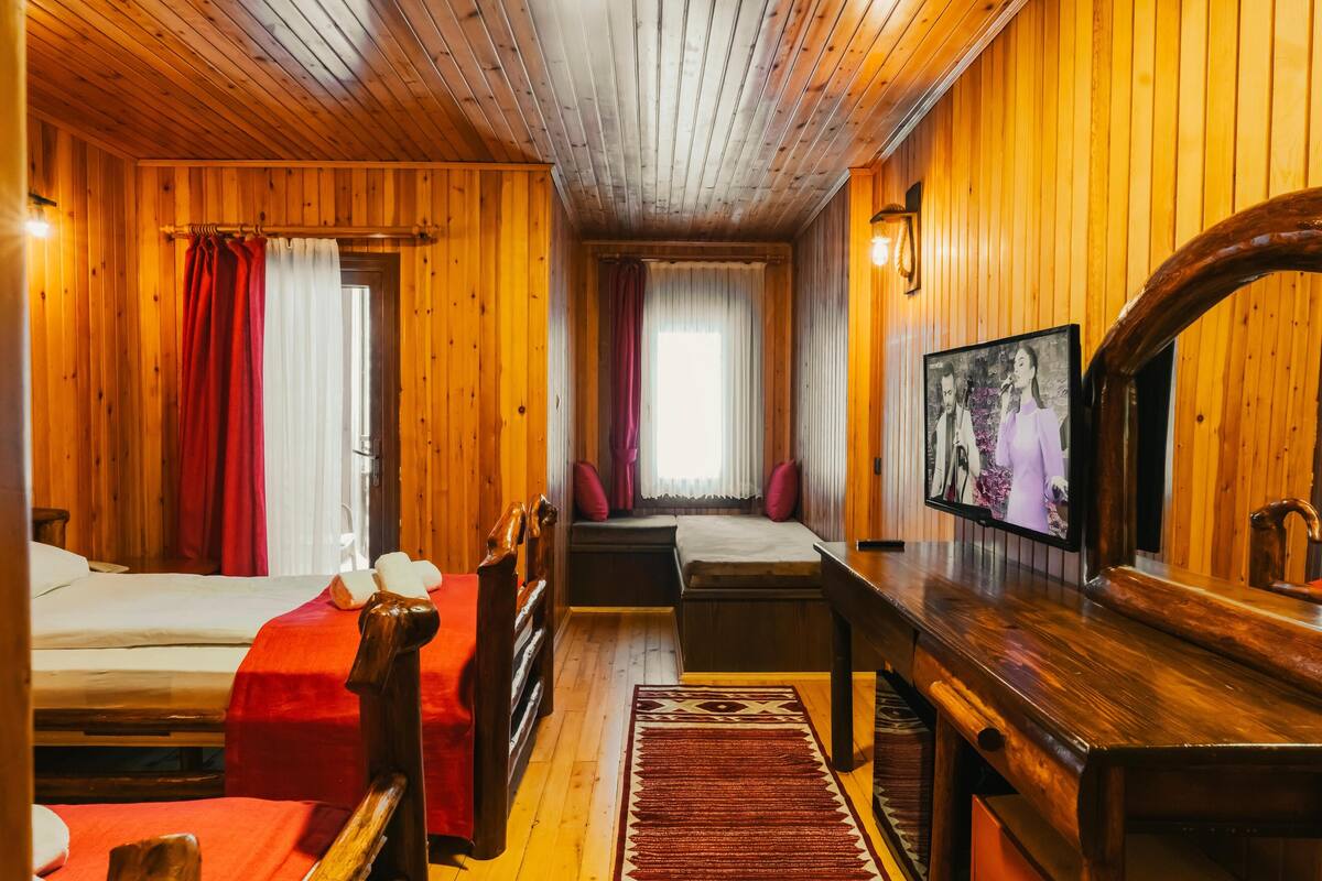 A wooden cabin room with two beds, a TV, and a window with curtains.