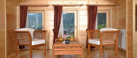 Une cabane en bois accueillante, avec deux chaises en bois, une petite table et une vue sur une forêt, visible à travers de grandes fenêtres.