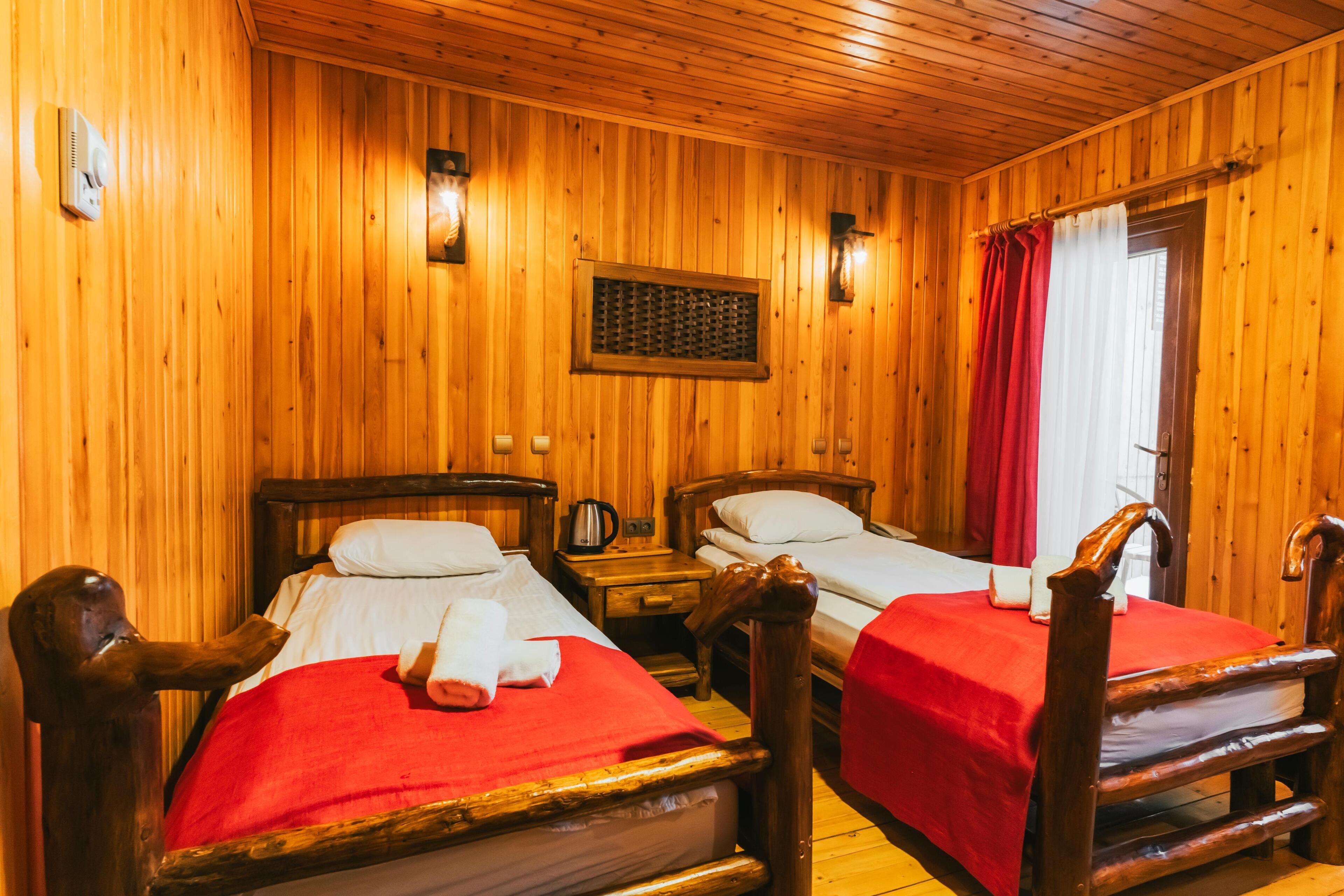 Una habitación de madera con dos camas, cada una con una colcha roja y almohadas blancas. Se observan cabeceros y mesitas de noche con lámparas de madera.