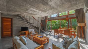Amplia sala de estar con techo de concreto, una escalera de madera, televisión y comedor con luz natural y vegetación visible a través de amplios ventanales.