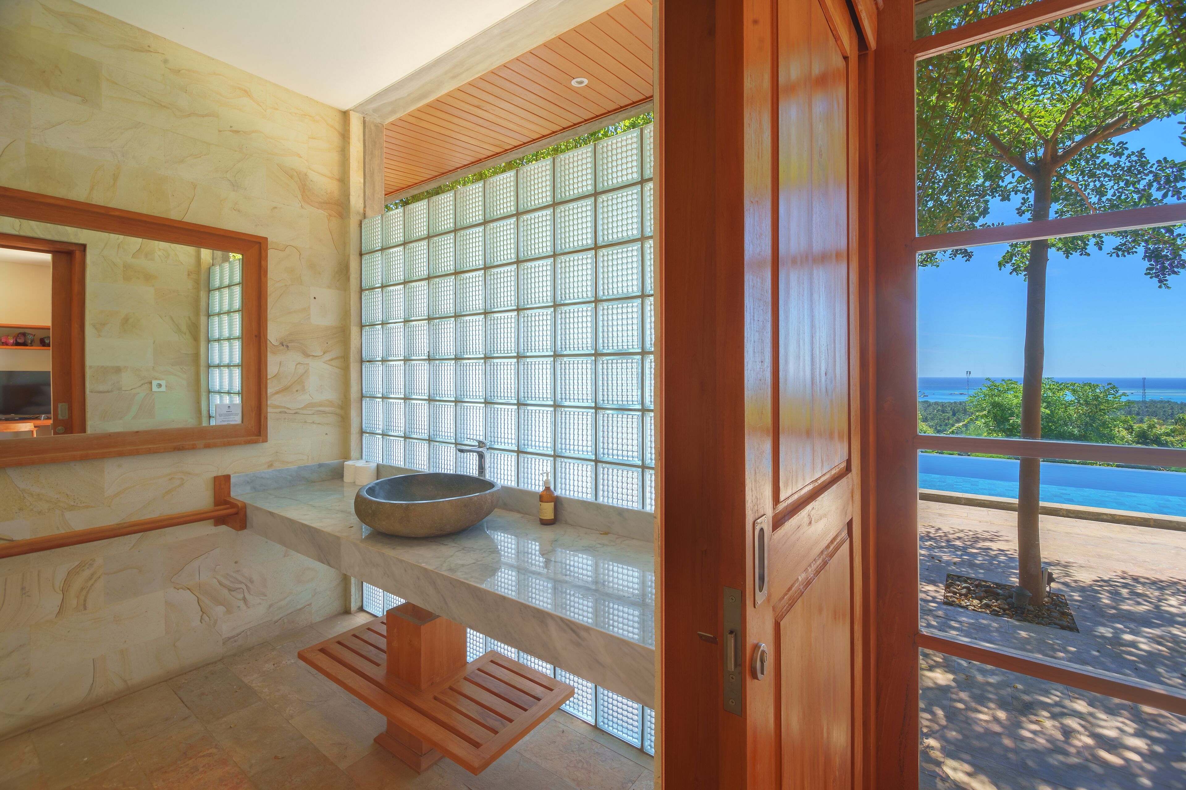 A modern bathroom with a large window, a wooden bench, and a stone sink.