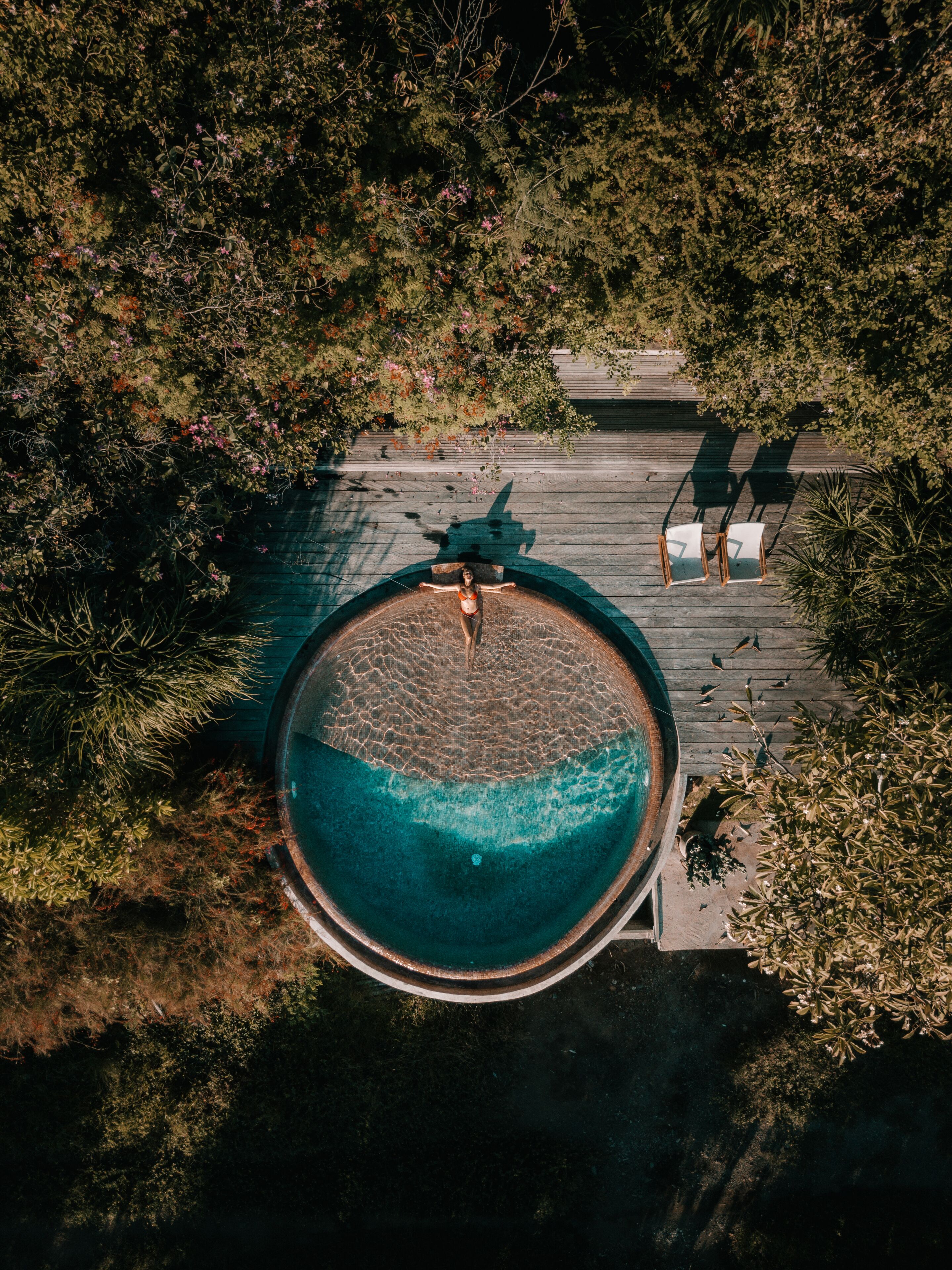An aerial view of a circular swimming pool surrounded by a wooden deck, with chairs and a bench for relaxation.