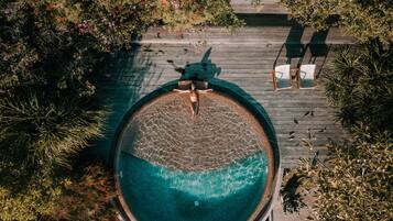 Vista aérea de una piscina circular rodeada por un muelle de madera, con sillas y un banco para relajarse.