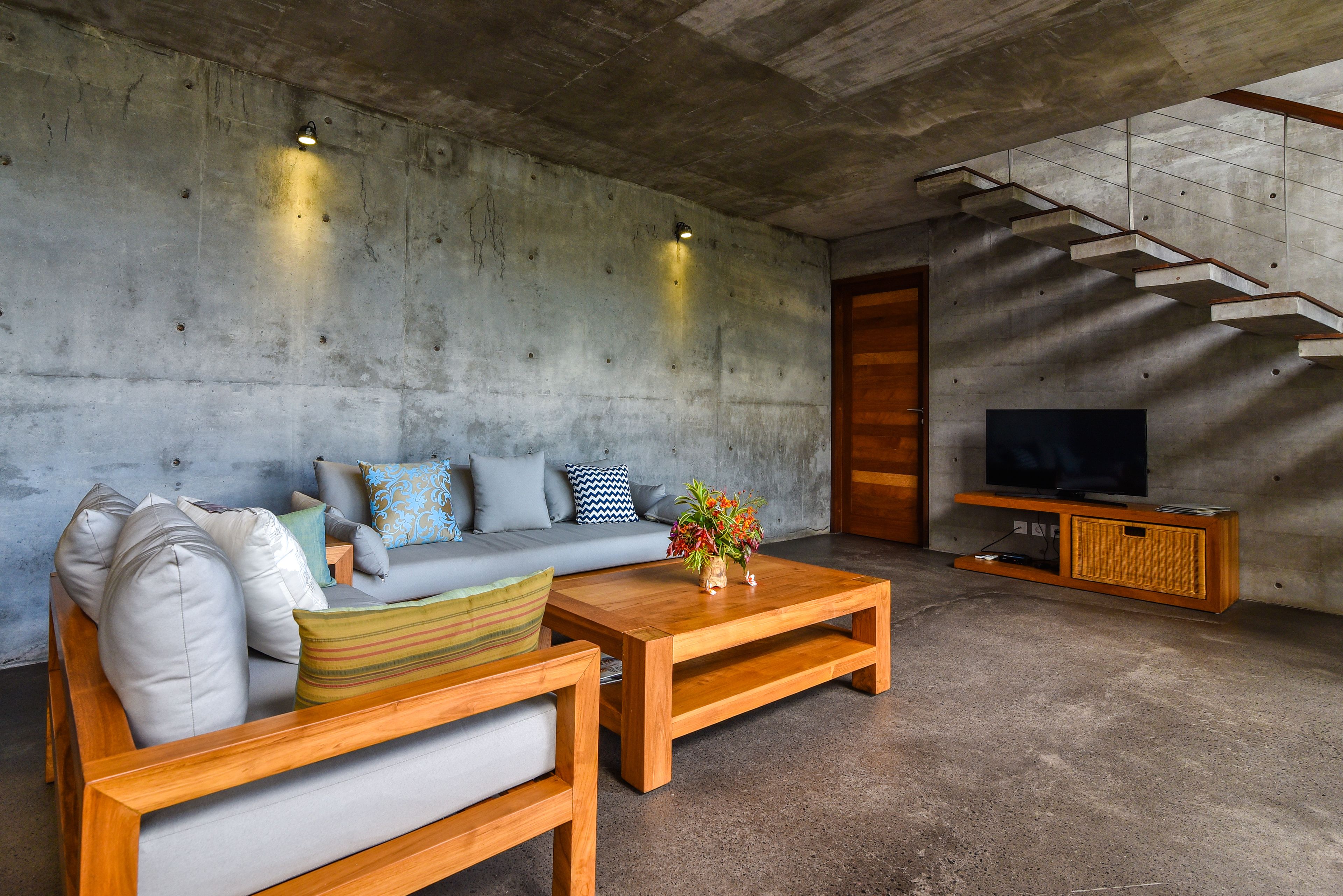 A modern living room with a concrete wall, a grey sofa, wooden coffee table, and a TV on a wooden stand.