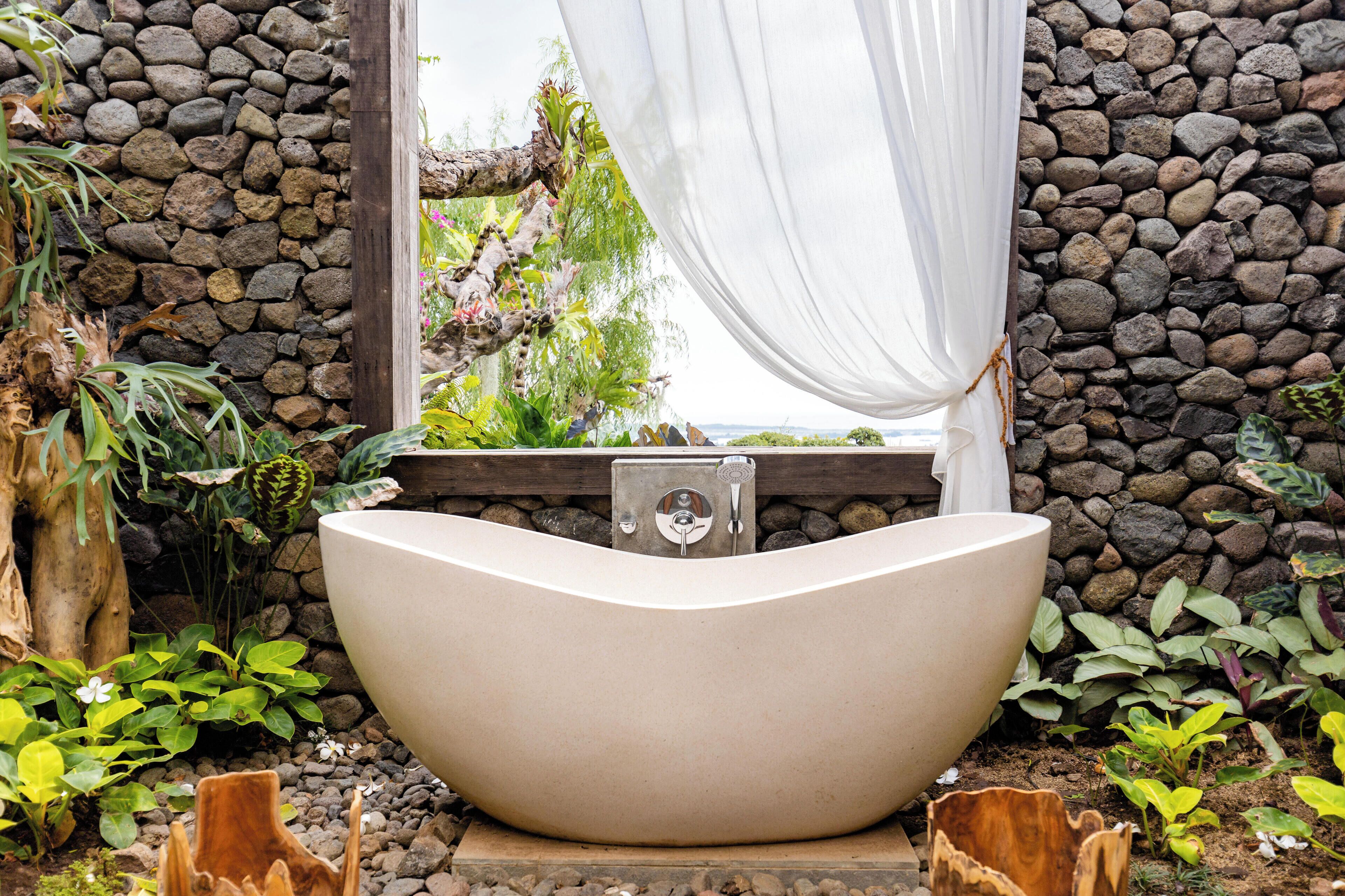 A freestanding bathtub in a natural setting with a stone wall backdrop and a window with curtains.