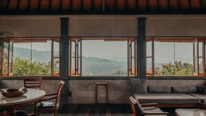 A room with a wooden floor, a round table with chairs, a sofa, and a window with an open shutter. - Sumberkima Hill Private Villa Retreat (Pemuteran)