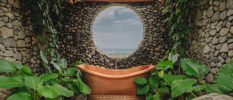 Une arche en pierre offrant une vue sur la mer, une baignoire en cuivre et une végétation luxuriante tout autour.