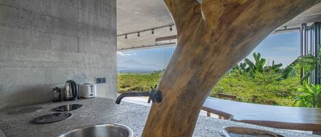 A kitchen with a large tree trunk centerpiece, a concrete countertop, stainless steel sink, and a view of greenery.