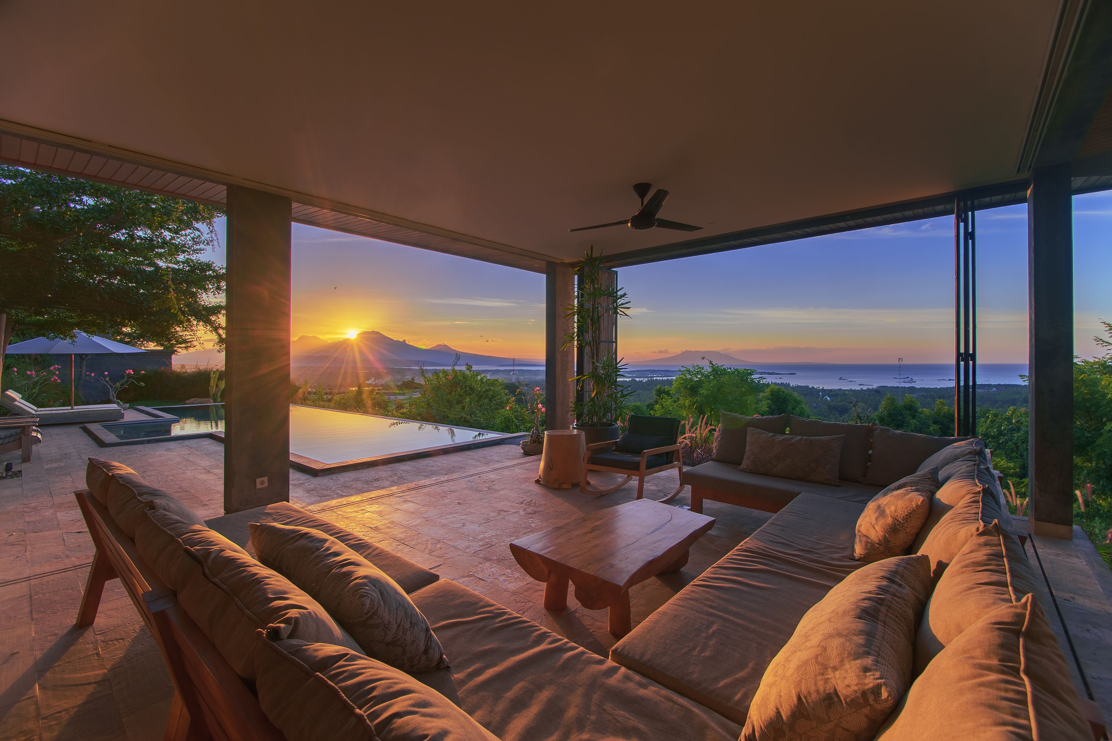 Ein großzügiger Außenbereich mit Blick auf den Sonnenuntergang über dem Meer, ein Pool und ein Essbereich mit Tisch und Stühlen.