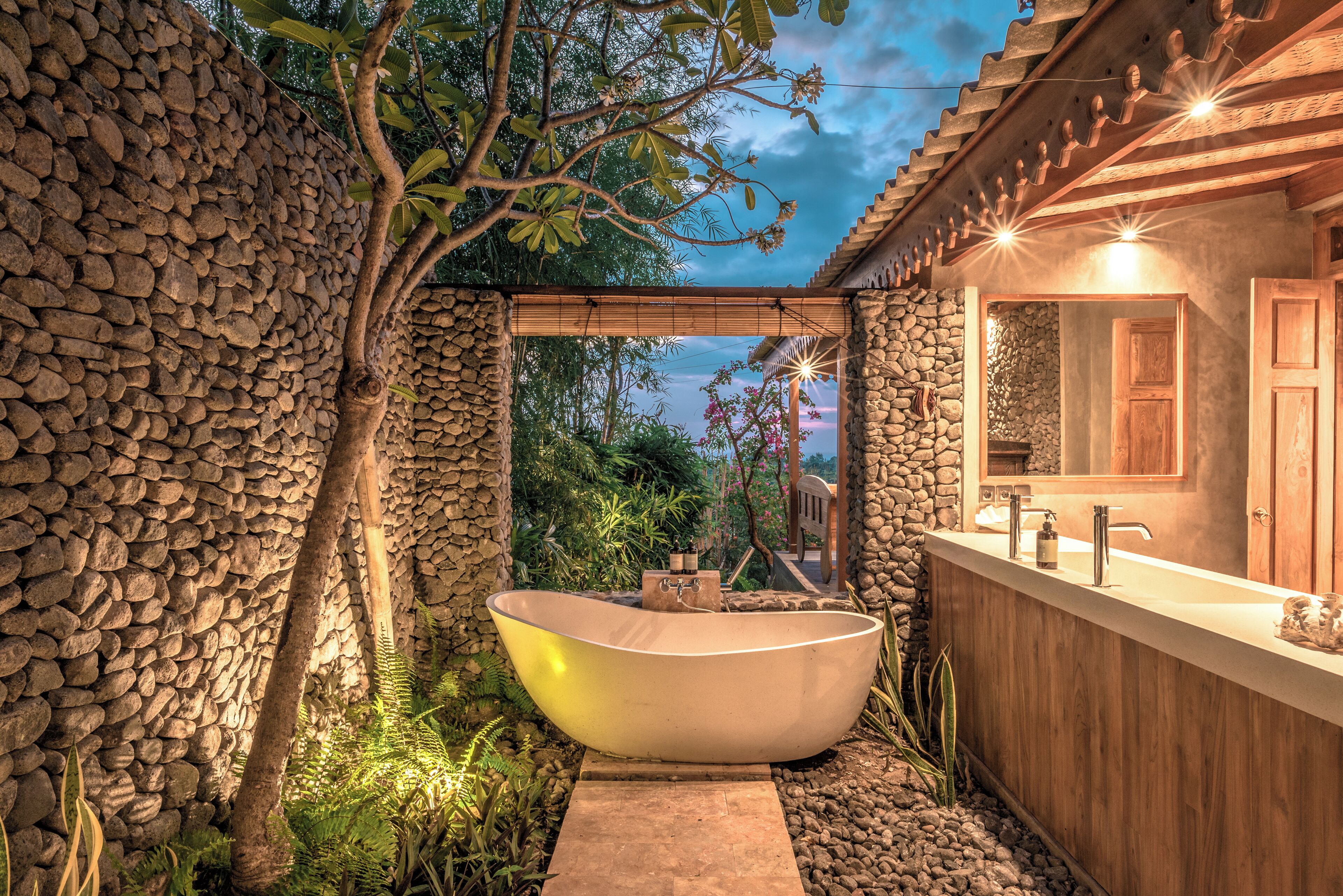 A stone-walled bathroom with a freestanding bathtub, a wooden vanity, and a stone archway.