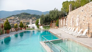 A pool with clear blue water, surrounded by white lounge chairs and umbrellas, with a stone wall and greenery in the background. - Maria Plomari Apartments (Lesvos)