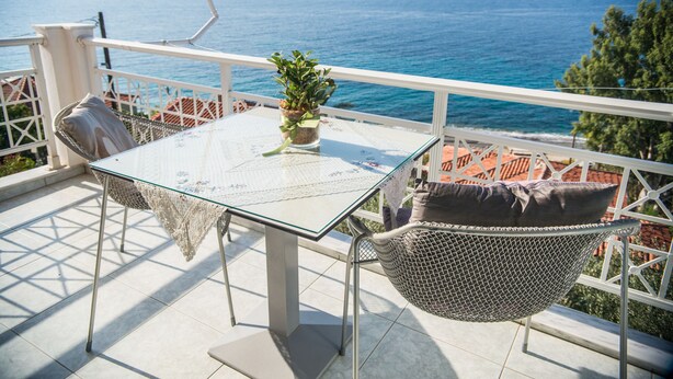 A glass table with a lace tablecloth, a potted plant, and wicker chairs on a balcony overlooking the sea. - Maria Plomari Apartments (Lesvos)