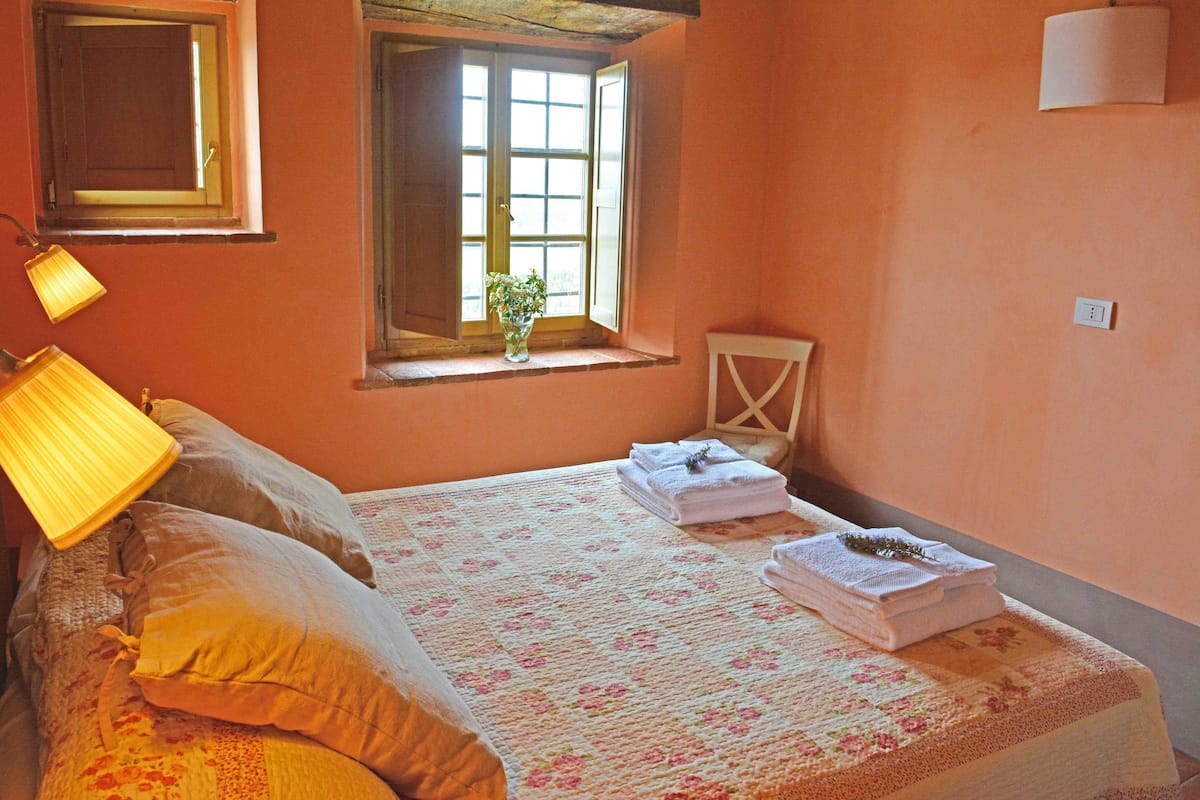 A bedroom with a bed, pillows, a bedside lamp, a window with a plant, and a chair.
