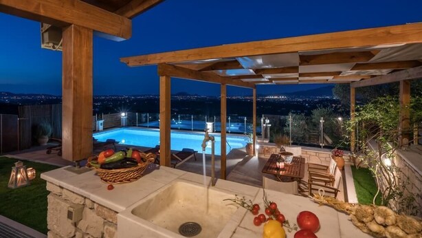 Un área de cocina al aire libre con un lavamanos de piedra, una cesta de frutas y vistas a una piscina por la noche.