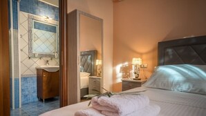 Habitación de hotel con cama, mesita de noche, espejo, tocador con lavabo y baño con azulejos azules.