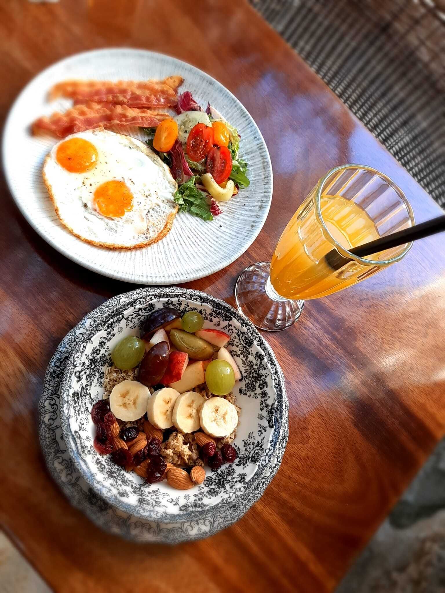 A breakfast setting with two plates, one with eggs and bacon, and another with fruit and nuts. There's a glass of orange juice with a straw.