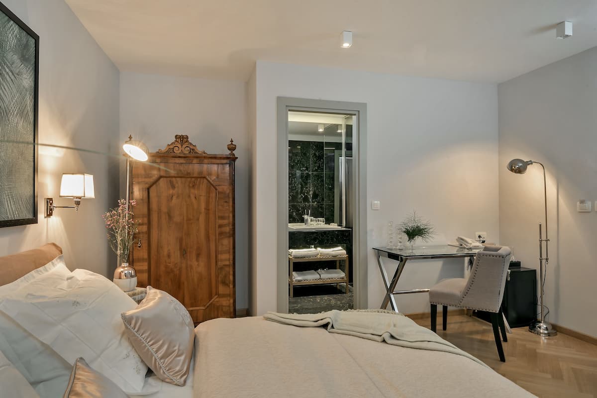 A bedroom with a bed, a wooden wardrobe, a desk with a chair, and a bathroom visible through a glass door.