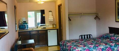 Quarto de hotel pequeno com cozinha compacta, cama com roupa de cama florida, escrivaninha com computador e uma cadeira.