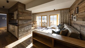 A modern interior with a wooden accent wall, a kitchen area, a seating area with a sofa and cushions, and large windows allowing natural light. - Alpbach Lodge Chalet Superior (Alpbach)