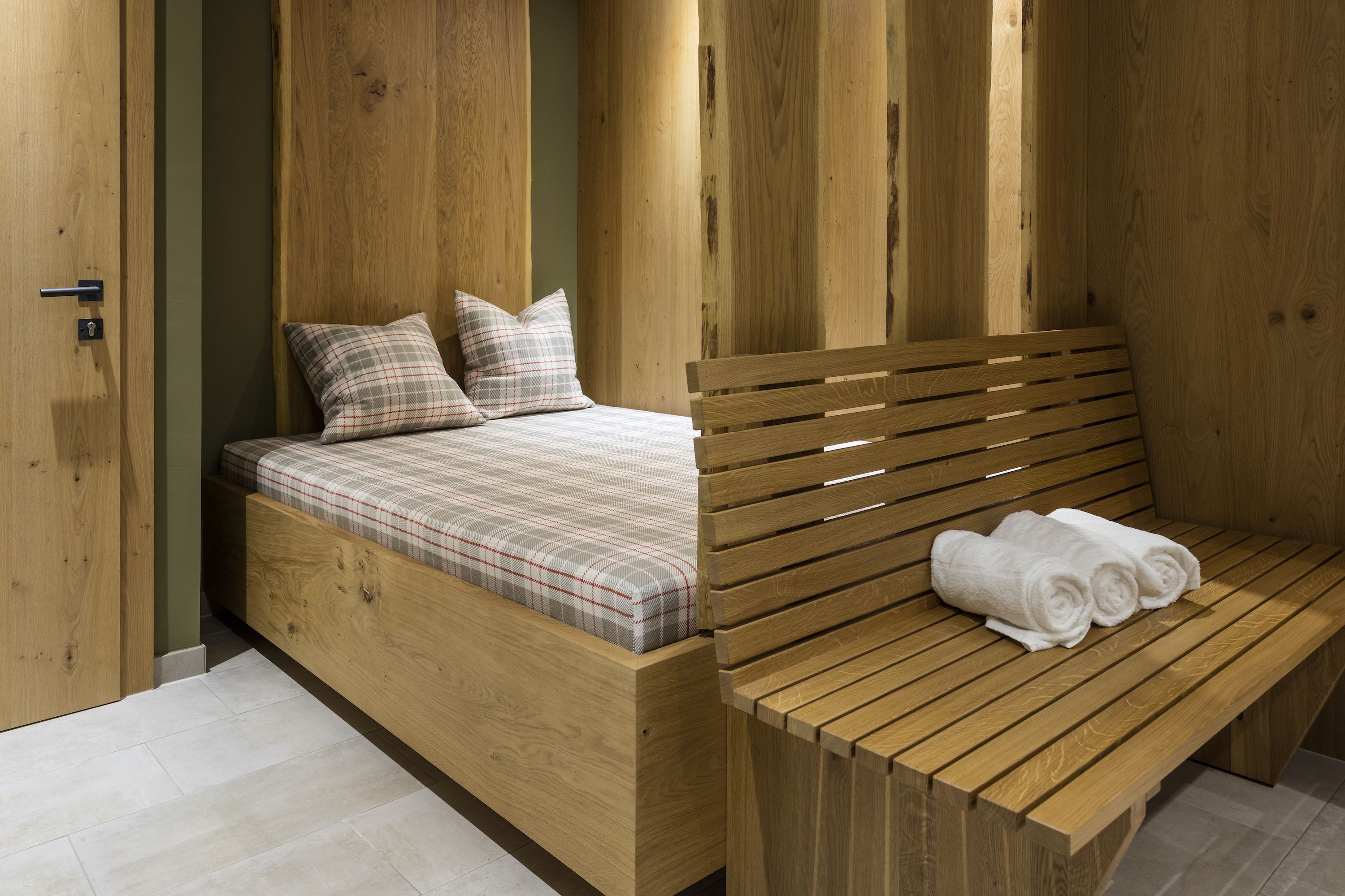 A wooden bed with a plaid mattress and pillows, a wooden bench with rolled towels, and a wooden door.
