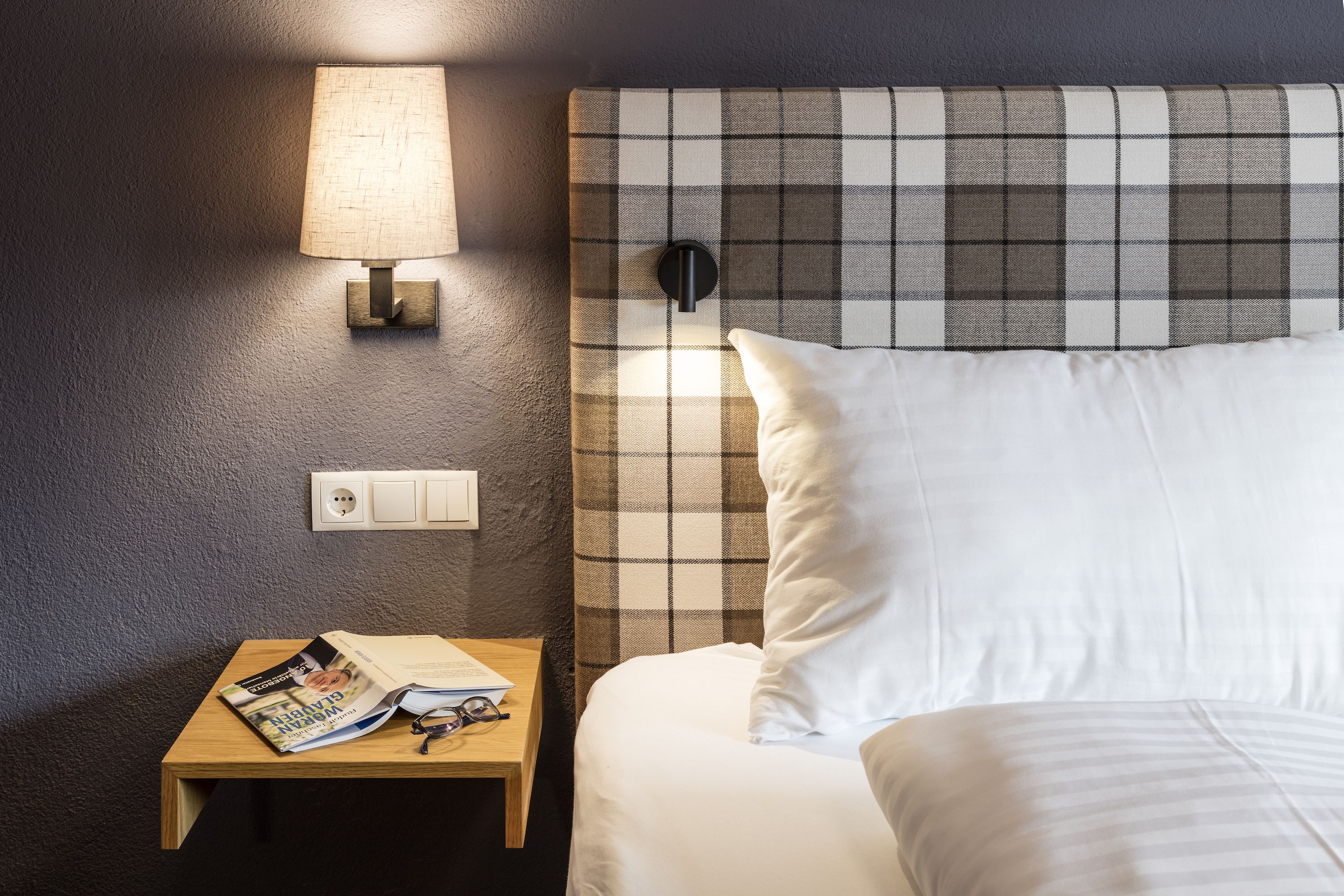 A neatly made bed with white linens, a plaid headboard, a bedside lamp, and a small wooden nightstand with a magazine and glasses.