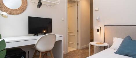 Ein Hotelzimmer mit einem Bett, einem Schreibtisch mit Computer, einem Sessel, einem Fernseher, einem kleinen Tisch mit Lampe und einer Tür zu einem anderen Raum.