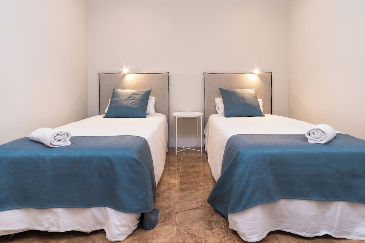 Habitación de hotel con dos camas, cada una con una colcha azul y almohadas blancas, y una pequeña mesita de noche blanca entre ellas.