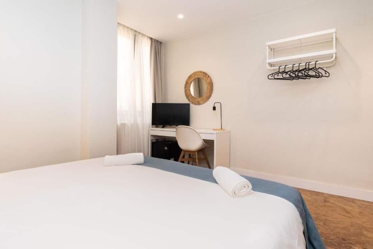 Una habitación de hotel con una cama, un escritorio con una silla, un televisor, un espejo fijado a la pared y un tendedero.