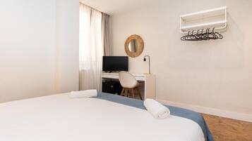 Una habitación de hotel con una cama, un escritorio con una silla, un televisor, un espejo fijado a la pared y un tendedero.
