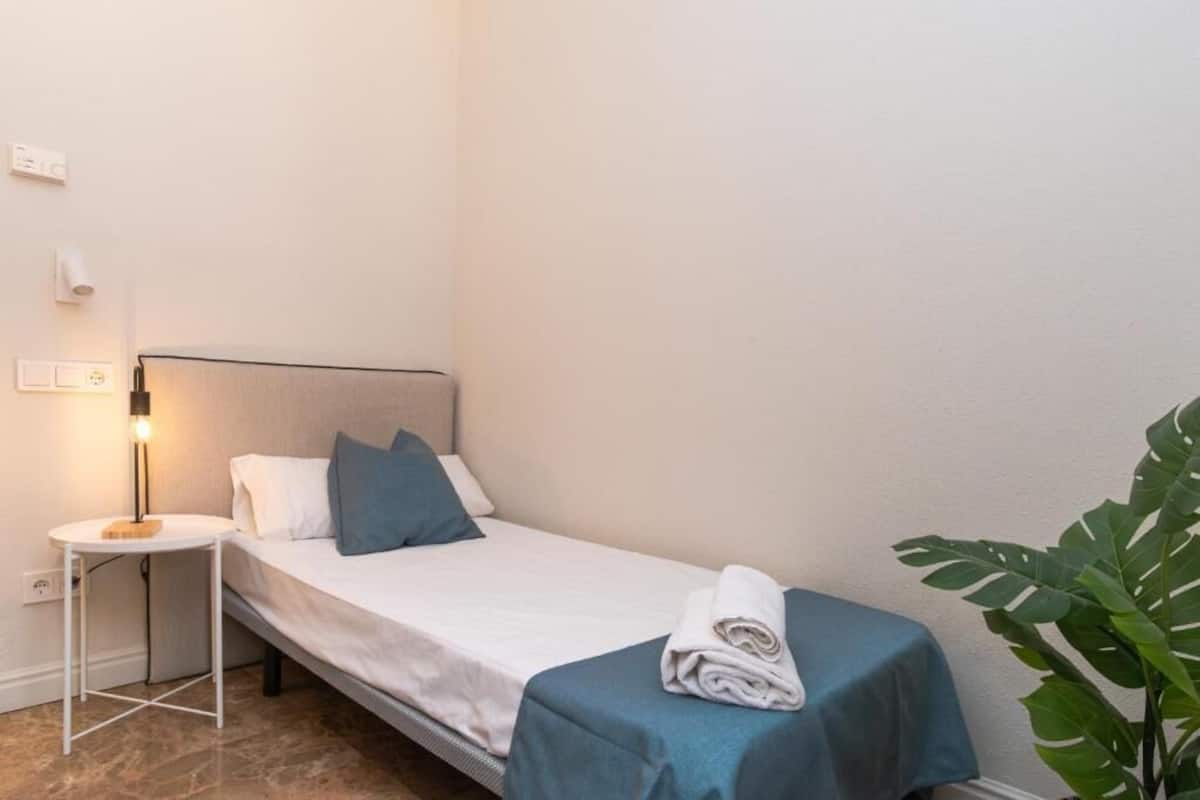 Una cama pequeña y bien arreglada con una colcha blanca y azul, una toalla enrollada al lado, una lámpara de noche y una planta en maceta en la esquina.