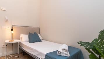 Ein kleines, ordentlich gemachtes Bett mit weißblauer Bettdecke, ein aufgerolltes Handtuch daneben, eine Nachttischlampe und ein Blumentopf in der Ecke.