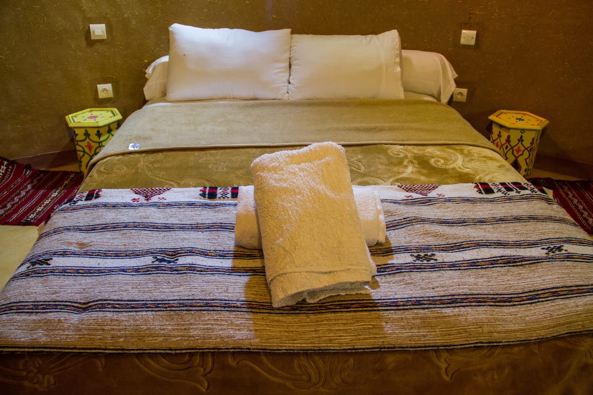 A bed with a patterned bedspread, two pillows, and a folded towel. There are two bedside tables with decorative bases.