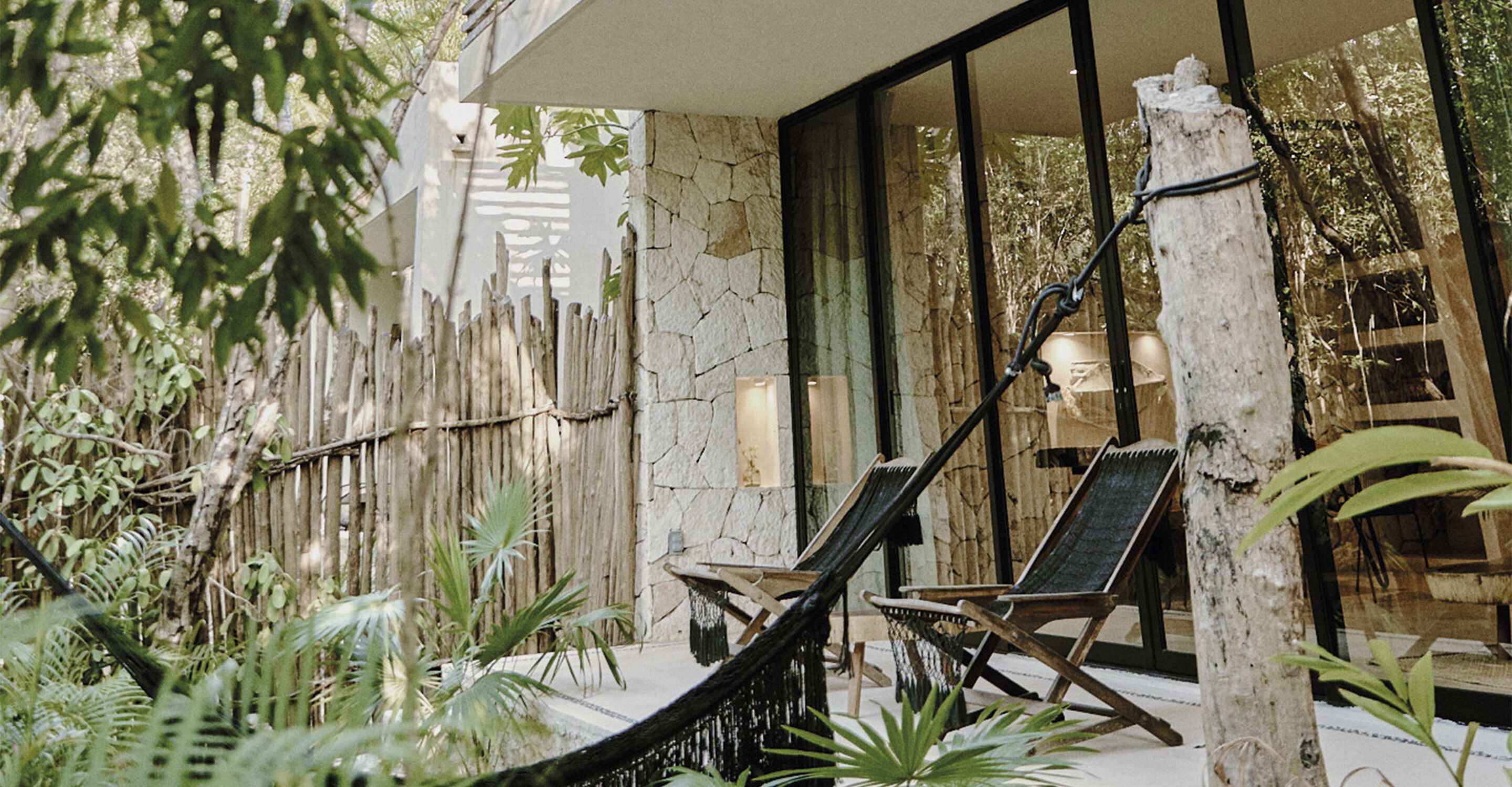 A hammock chair and a lounge chair in a tropical setting with a bamboo fence and lush greenery.