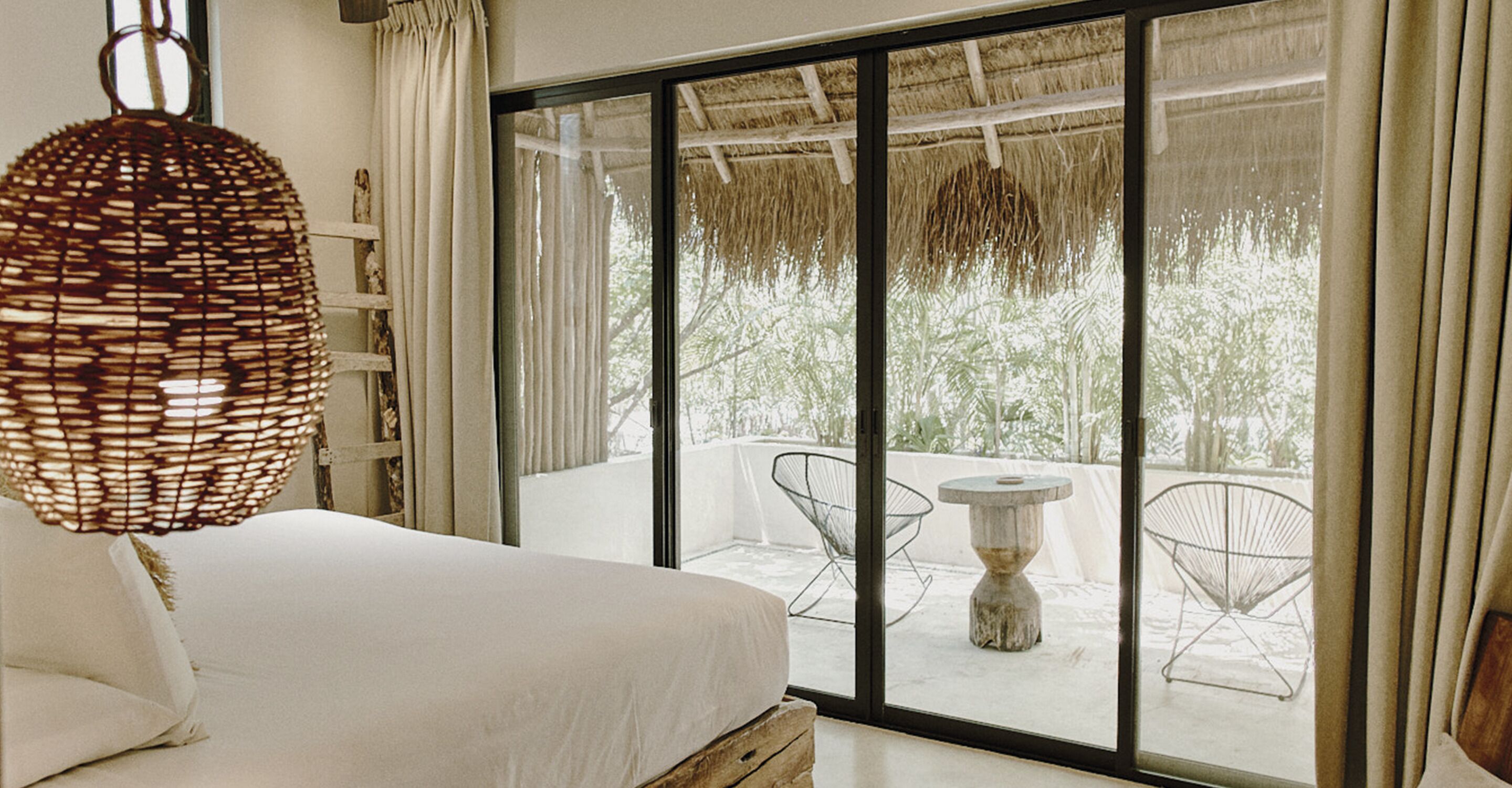 A bedroom with a large window, a wicker hanging light fixture, and a view of an outdoor area with chairs and a table.