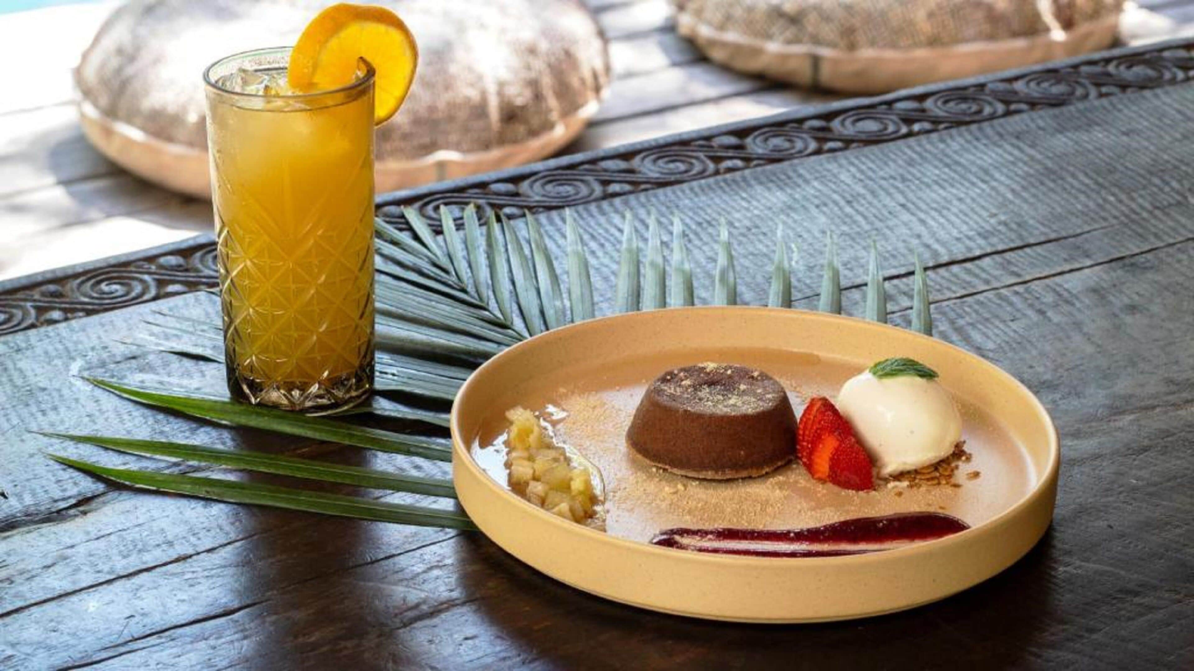 En desserttallerken med en chokolademousse, en is kugle, og en jordbær, ledsaget af et glas gul drik med et appelsinstykke.