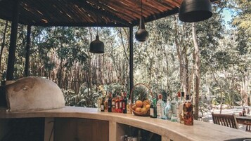 Area bar all'aperto con un bancone in legno rustico, luci a sospensione e una selezione di bottiglie e frutta.