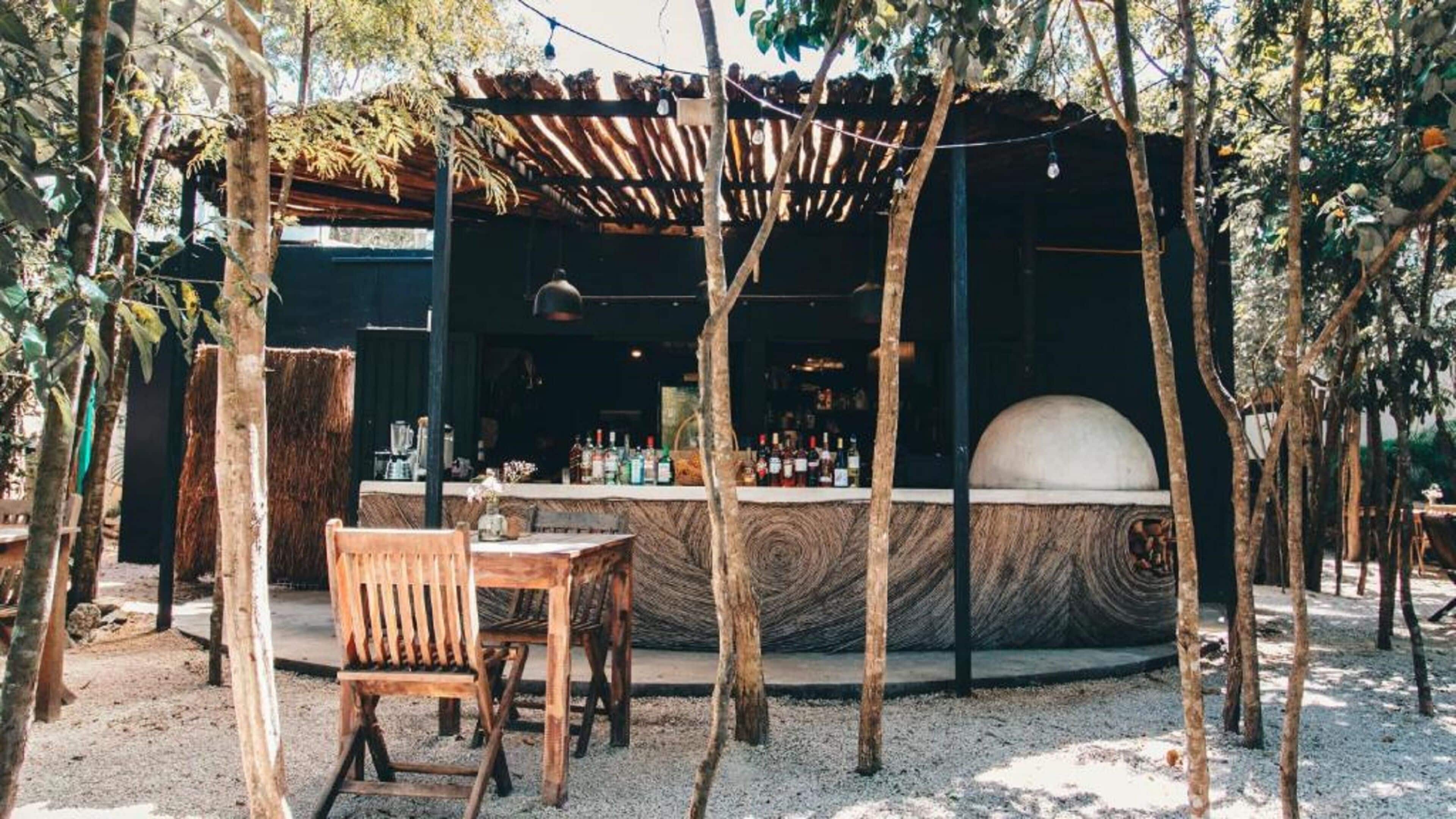 Een terras met een houten tafel en stoel, een bar met een toonbank en flessen, en een grote koepelstructuur.